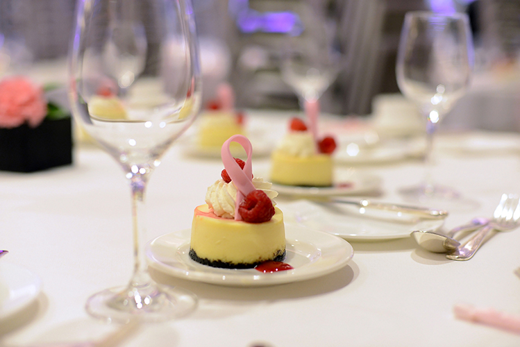 A small cheesecake topped with whipped cream, raspberries, and a pink sugar ribbon sits on a white plate. The table is set elegantly with empty wine glasses and silverware.