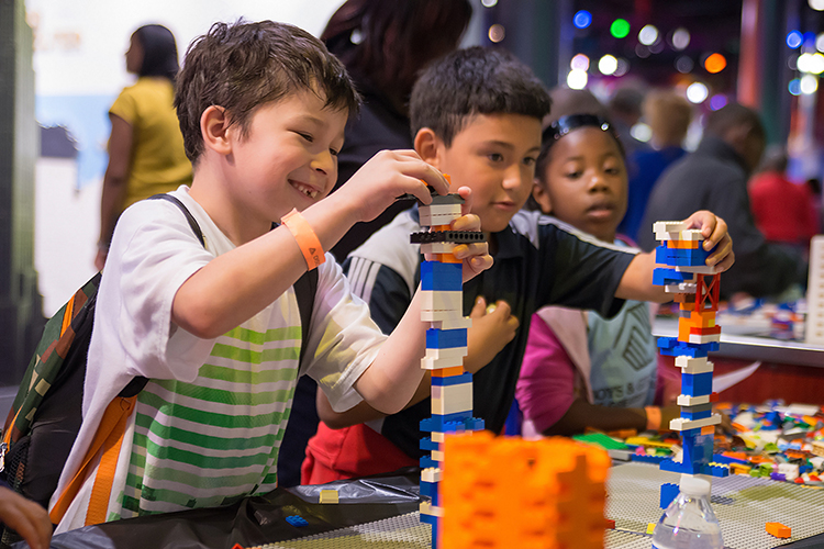 Children are building colorful structures with LEGO bricks at a table in a busy activity center. The atmosphere is lively with people in the background, and the kids appear focused and engaged in their creative play.