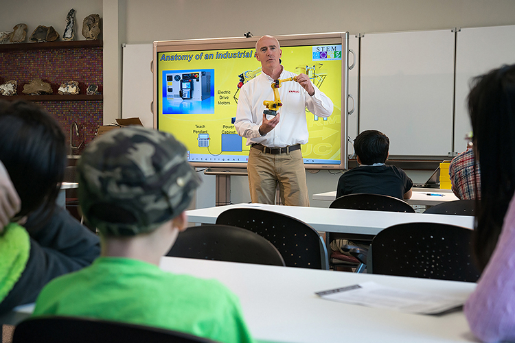 A man wearing a white shirt is giving a presentation in a classroom, holding a model. Several children are seated at tables, watching attentively. A large interactive display board shows diagrams and images,