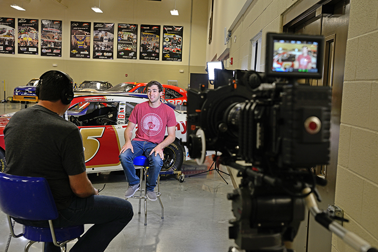 A young man in a red shirt is being interviewed in front of a race car inside a garage. The interviewer sits opposite him, wearing headphones. A camera is focused on the young man.