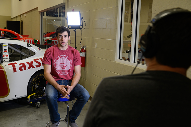 A young man in a red shirt is being interviewed in front of a race car inside a garage. The interviewer sits opposite him, wearing headphones.