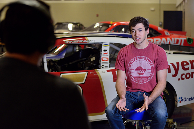 A young man in a red shirt is being interviewed in front of a race car inside a garage. The interviewer sits opposite him, wearing headphones.