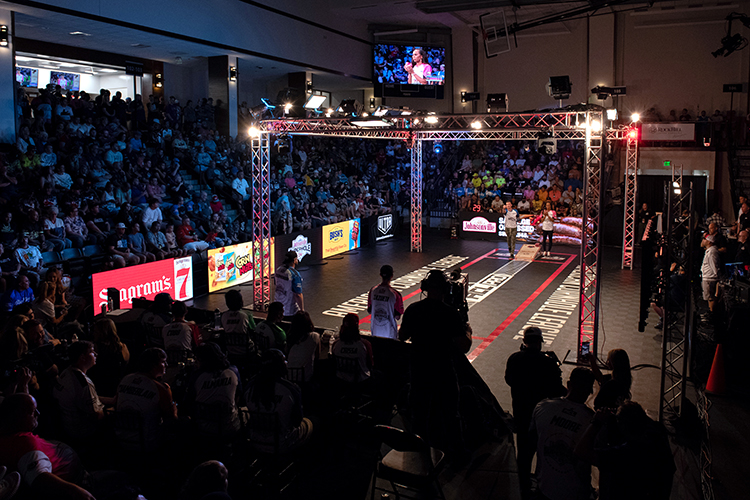A lively indoor sports event with a square arena surrounded by an enthusiastic audience. Large screens and bright lights illuminate the venue.