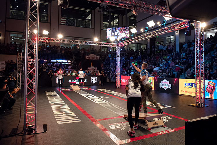 Two people compete in a cornhole match under bright lights in an indoor arena. Spectators fill the stands in the background. The floor features various logos, and two large screens display the game.
