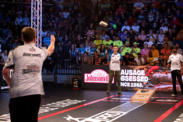 A lively cornhole tournament with players tossing bean bags toward a board. The audience watches from the stands, and a digital scoreboard displays the score. A large Johnsonville banner is visible in the background.