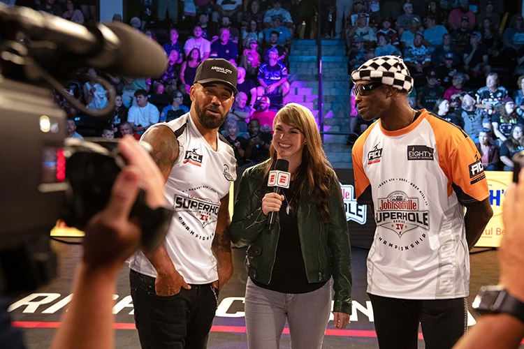 A woman holding a microphone interviews two men wearing sports jerseys at a live event. They are standing in front of a lively audience. A camera is visible in the foreground capturing the scene.