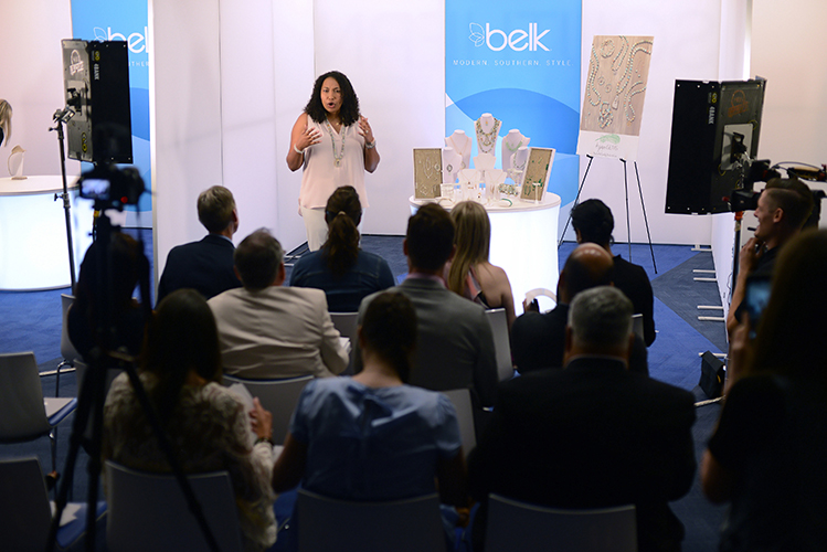 A presenter stands in front of an audience, speaking at designer showcase. The audience is seated, and several cameras are set up to record the presentation.