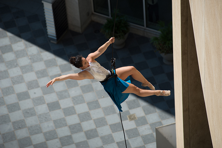 Aerial performer suspended on a rope, wearing a blue skirt and white top, pushes off from the face of a building over a tiled plaza below.