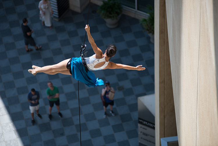 Aerial performer suspended on a rope, wearing a blue skirt and white top, extends her arms and legs horizontally outside a building. Several onlookers observe from below.