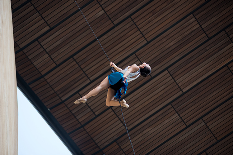 The view looking up of an aerial performer suspended on a rope, wearing a blue skirt and white top, leaps out from the edge of a building.  A wooden ceiling is seen in the background.