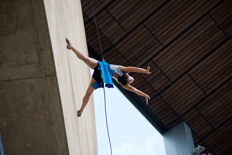 A view looking up at an aerial performer suspended on a rope, wearing a blue skirt and white top, extends her arms and legs posed on the side of a building.