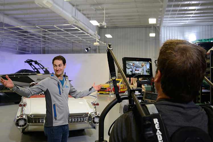 A person in a gray jacket stands with arms outstretched, facing a video camera setup in a garage studio. In the background, theres a classic white car and a parked boat.