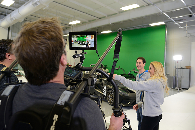 A crew member holds a video camera and display screen filming a man and woman in a studio with a luxury sports car in the background.