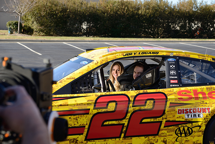 A man and woman sit inside a NASCAR racecar parked in an empty lot while a camera crew films them.