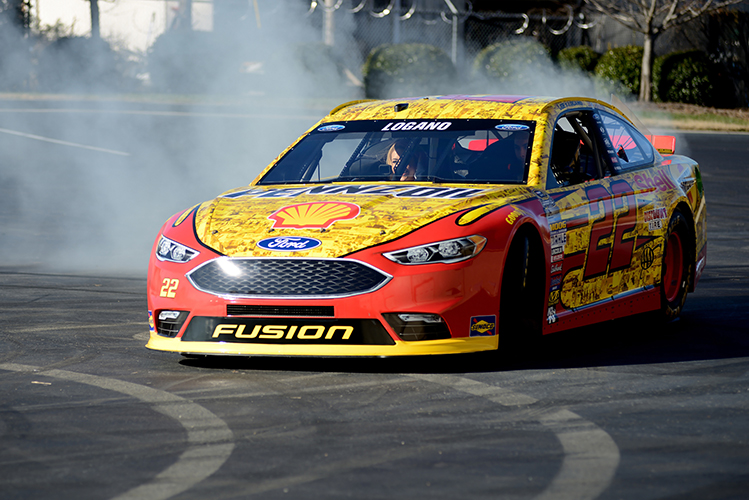 Smoke billows from behind a yellow and red NASCAR racecar as it skids around an empty parking lot. A woman is seen in the car laughing.