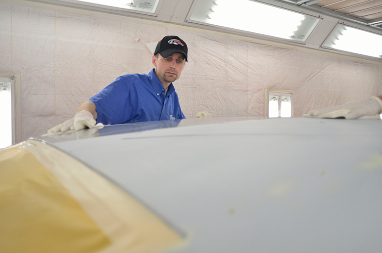 A person wearing a blue shirt and black cap cleans a car’s surface before painting. He is in a will lit painting booth.