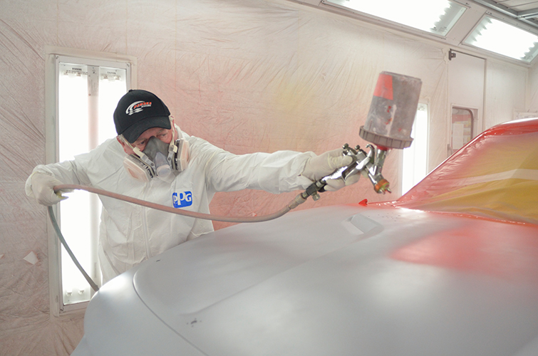 A person wearing protective gear and a respirator is spray painting a car in a workshop with a spray gun.