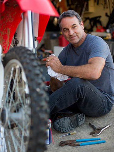 A man sits on the floor of a garage, holding a cloth and surrounded by tools, while smiling. He appears to be working on a dirt bike, which is partially visible in the foreground.