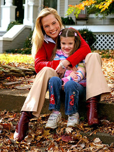 A smiling woman in a red sweater and beige pants sits on steps with a young girl in colorful clothes, surrounded by fallen autumn leaves, in front of a house. They both look happy and cozy.