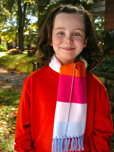 A smiling child with curly hair wears a bright red sweater and a colorful scarf with pink, orange, and blue stripes. She stands outside in a leafy area with trees and sunlight filtering through