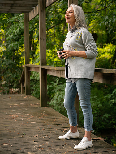 An elderly woman with gray hair stands on a wooden boardwalk holding binoculars and smiling as she looks into the distance. She is wearing a gray sweater with floral designs, jeans, and white sneakers.