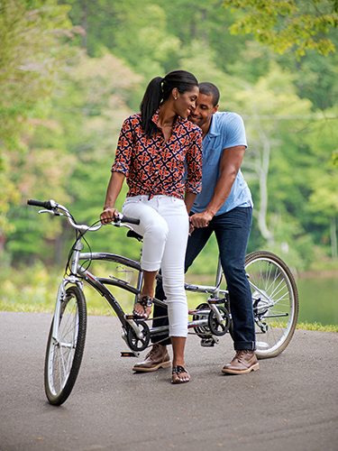 A couple enjoys a tandem bicycle ride on a paved path surrounded by lush greenery. The woman smiles as she sits on the bike, while the man leans in and gently holds her shoulders.