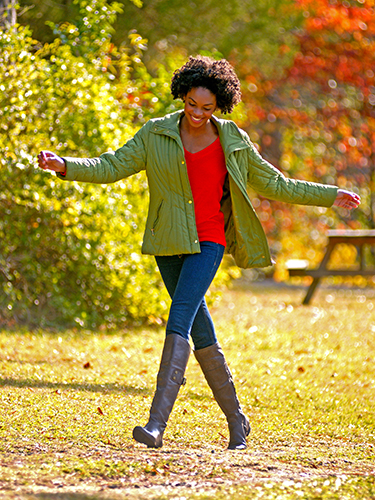 A woman joyfully walks outdoors on a sunny day, wearing a green jacket, red shirt, jeans, and tall gray boots. The surroundings are filled with lush greenery and trees with autumn leaves.