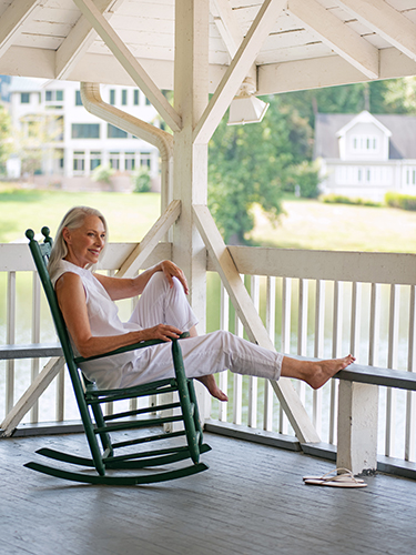 A woman with gray hair relaxes in a dark green rocking chair on a porch, wearing white clothing. She has one leg up on the chair, looking content. The background features a calm lake and houses surrounded by trees.