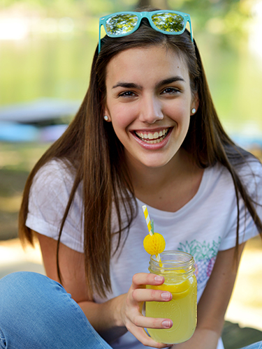 A woman with long brown hair smiles while holding a jar of lemonade with a straw outdoors. She is wearing a white t-shirt and blue sunglasses on her head. The background is a blurred natural setting.