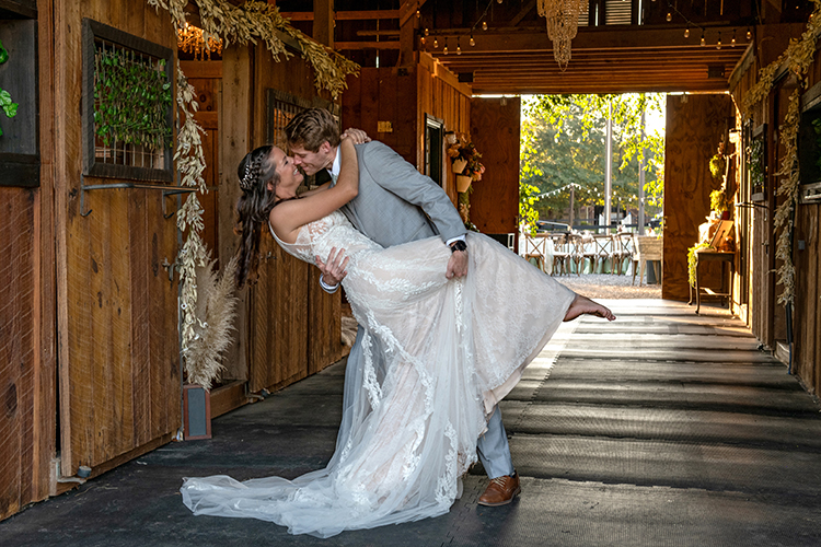 A groom in a light gray suit romantically dips and kisses his bride, who is wearing a white lace gown, inside a rustic barn decorated with dried flowers. Sunlight filters through the open doorway, illuminating the couple.
