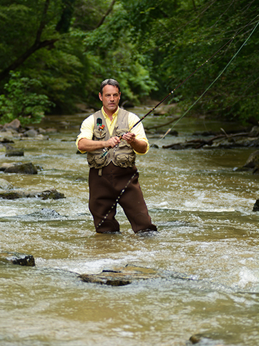 A man wearing waders and a vest stands in a flowing river while fly fishing, surrounded by lush green forest.