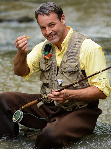 A man crouches by a river, smiling while holding a fly fishing rod. Hes wearing a beige fishing vest over a yellow shirt and brown waders, and appears to be enjoying the outdoor activity.