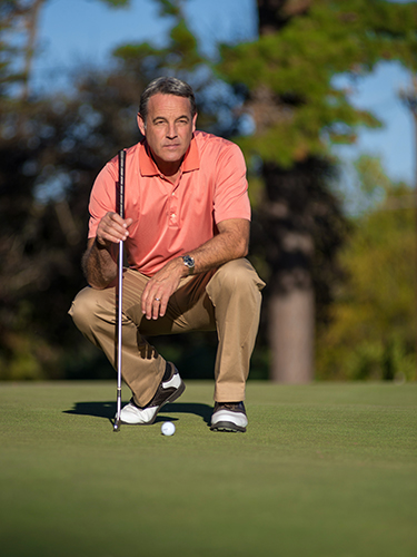 A man in a coral polo shirt and khaki pants crouches on a golf course, intently focusing on the placement of a golf ball. He holds a putter and is surrounded by blurred trees in the backgroundgreen.