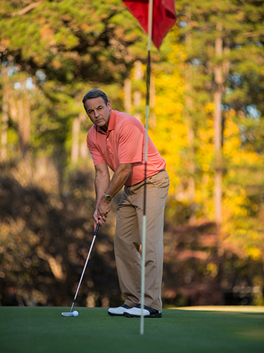 A man in a pink polo shirt and beige pants is preparing to putt on a golf course. The scene is set against a background of trees with autumn foliage, and a red flag is visible near the hole.