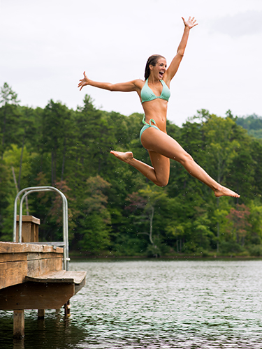 A woman in a green bikini jumps joyfully into a lake from a wooden dock. The background features lush green trees and a calm water surface, capturing a lively summer moment.