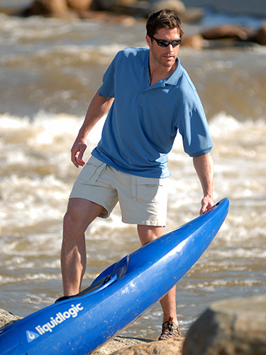 A man wearing sunglasses, a blue polo shirt, and beige shorts is standing on rocky terrain by a river. He is holding the edge of a blue kayak, looking away from the water.