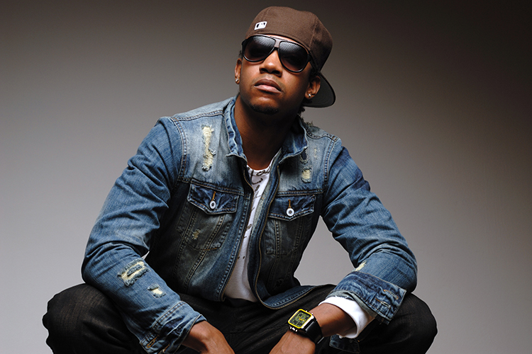 A person wearing a denim jacket, sunglasses, and a brown cap sits against a neutral background. They have their hands on their knees and are also wearing a watch on their left wrist.