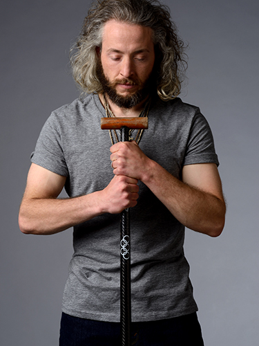 A man with long curly hair and a beard wearing a gray shirt holds a canoe paddle centered on his chest with both hands. The five ring logo of the Olympics is printed on the paddle.