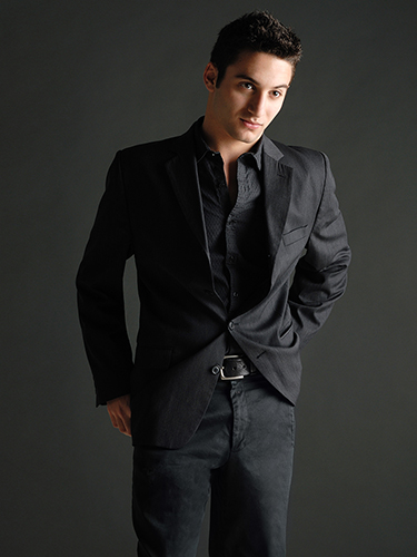 A man stands confidently against a dark background, wearing a black suit jacket and pants with a black shirt. His hands are in his pockets, and he gazes slightly to the side with a composed expression.