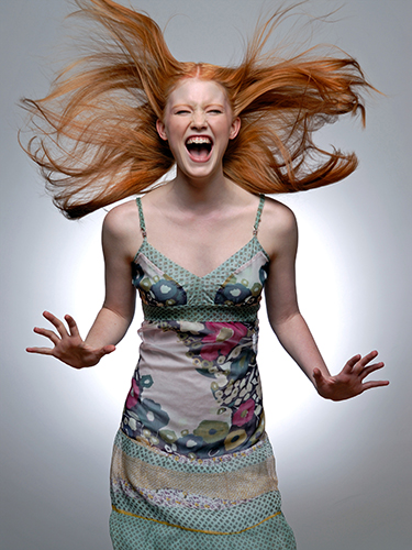 Portrait of a woman with red hair wearing a sleeveless, patterned dress. Her hair flows upwards as she shouts in elation against a plain white background.