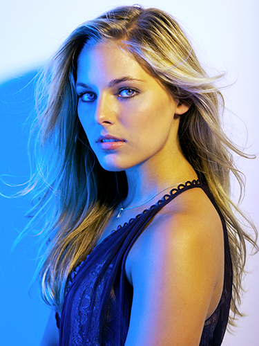 A woman with long blonde hair cast in cool blue light on the left and warm yellow light on the right. Her hair is wavey and she is wearing a a sleeveless black top and a necklace.