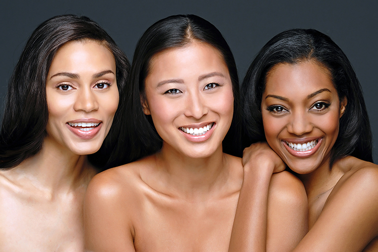 Three women smiling, positioned closely with bare shoulders, set against a dark background. They exhibit joyful expressions, with their different skin tones and straight, dark hair creating a harmonious and vibrant scene.