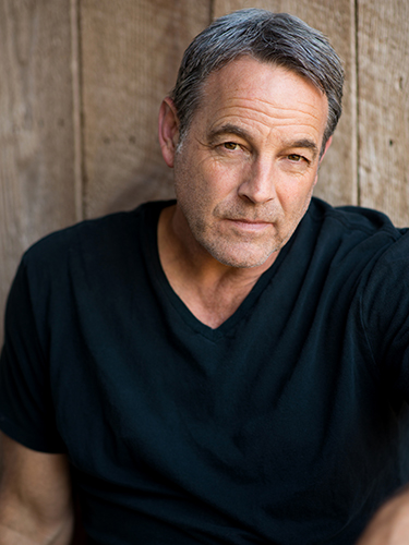 A middle-aged man with short gray hair and wearing a black T-shirt is sitting in front of a wooden background, looking at the camera. He has a slight smile with a relaxed expression.