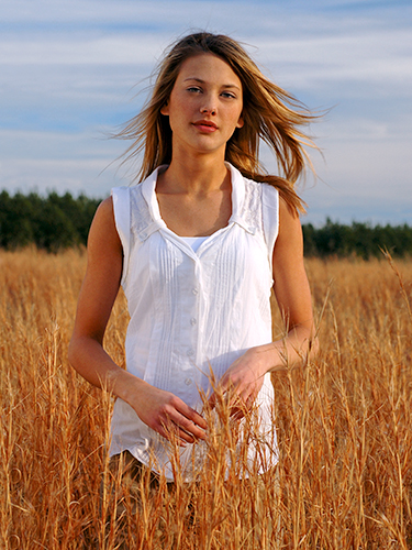 A woman with long hair stands in a field of tall, golden grass. They are wearing a white sleeveless blouse. The sky is blue with some clouds, and trees line the horizon in the background.