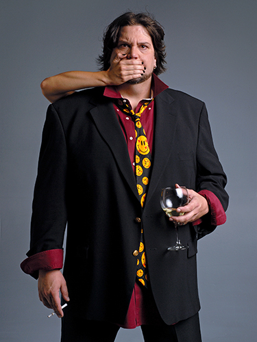 A man in a black blazer and a smiley face tie holds a wine glass in one hand and a cigarette in the other. A hand covers his mouth from behind. The background is a plain gray.