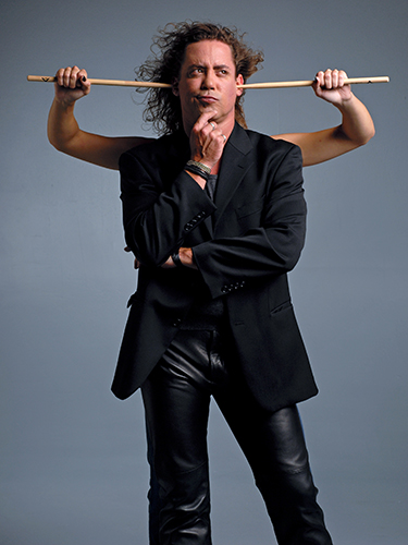 A man in a black suit and leather pants stands with a thoughtful expression. Behind him, another set of arms holds drumsticks in his ears against a gray background.