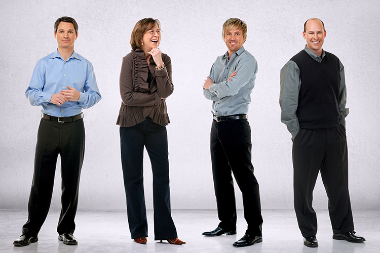 Four people standing against a plain background. Three men are wearing smart-casual outfits: shirts and pants; one also has a sweater vest. A woman wears a blouse and pants. They appear relaxed and are smiling.