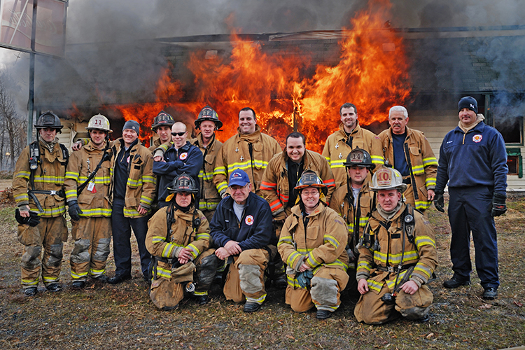 A group of firefighters in full gear poses for a photo in front of a raging fire. The flames are intense, and smoke rises from a building structure behind them.