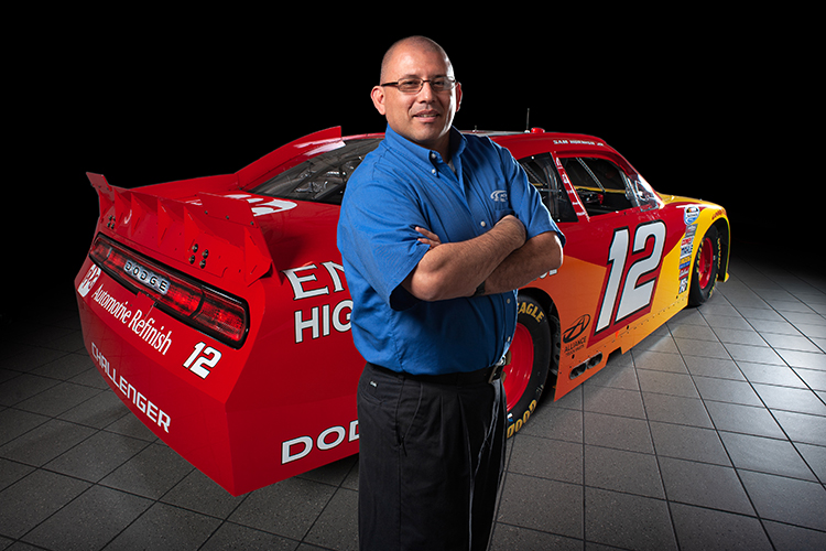 A man in a blue shirt stands confidently with arms crossed in front of a red and yellow racing car on a tiled floor. The car features the number 12 and various sponsor logos. The background is black, highlighting the subject and vehicle.