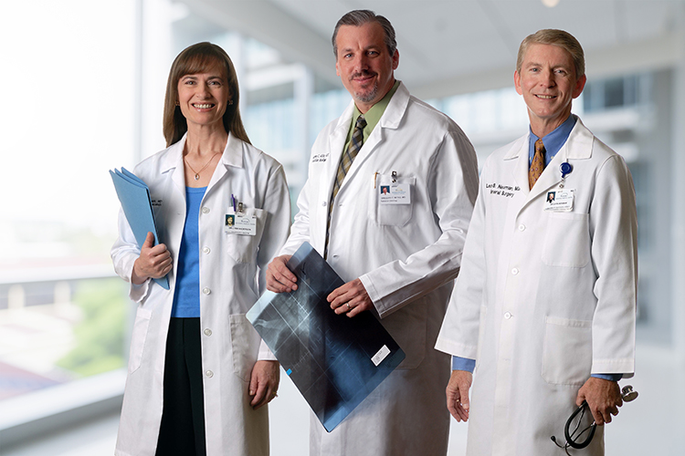 Three doctors in white coats stand in a bright, modern hospital corridor. Two men and a woman, all holding medical documents, pose confidently. They wear professional attire and ID badges, suggesting they are experienced medical professionals.